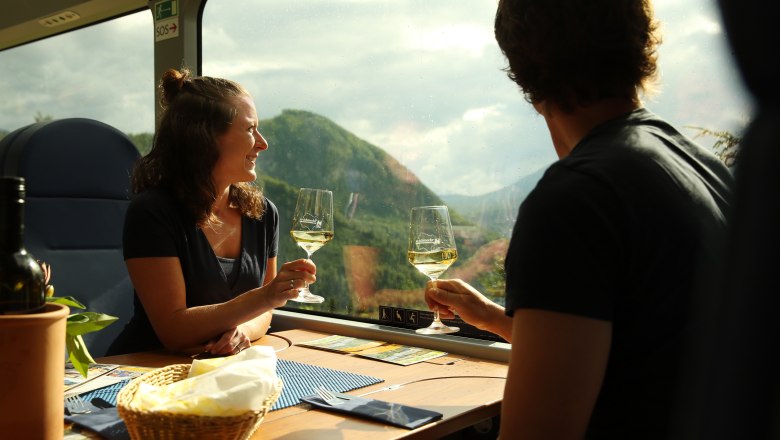 Feldversuche in der Mariazellerbahn und im Naturpark Ötscher:Tormäuer, © NÖVOG/Weinfranz Zwei Personen sitzen an einem Tisch im Zug und stoßen mit Weingläsern an, im Hintergrund sind Berge durch das Fenster zu sehen.