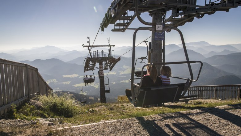 Lift ride to the Gemeindealpe Mitterbach, © Robert Herbst Two people on a chair lift with a mountain landscape in the background.