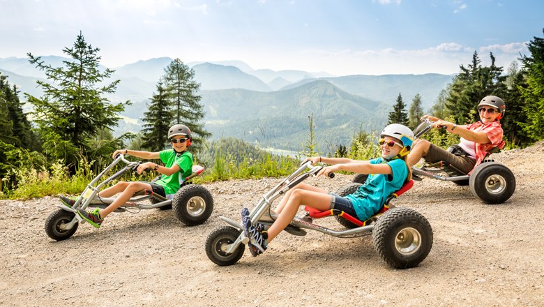 Children and adults alike love the fast-paced ride, © Lindmoser Three people ride down a mountain on mountain carts, surrounded by a mountain backdrop.