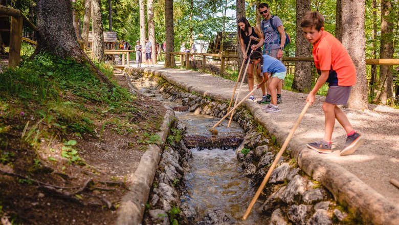 Holzknechtland Bürgeralpe, © Bürgeralpe/Dellinger Familie spielt mit Stöcken in einem Wasserkanal im Wald.