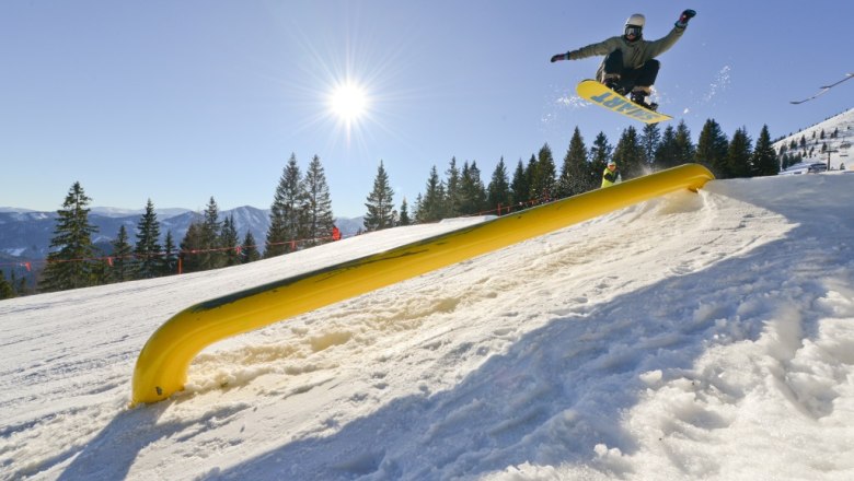 In winter, the Gemeindealpe is the destination for winter sports enthusiasts, © Bergbahnen Mitterbach Snowboarder jumping over a yellow obstacle on a snowy slope in the sunshine.