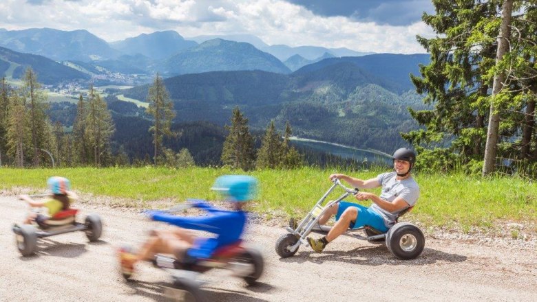 The mountain carts take you swiftly down into the valley, © Bergbahnen Mitterbach People ride down the mountain on mountain carts, an impressive mountain landscape in the background.