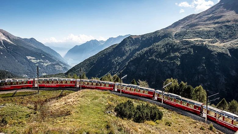 Ein rot-weißer Panoramazug fährt auf einer kurvigen Bahnstrecke durch eine eindrucksvolle alpine Berglandschaft mit weitem Blick ins Tal.