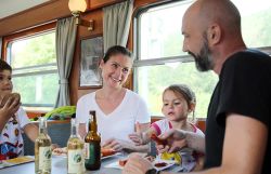 Eine vierköpfige Familie sitzt an einem Tisch in einem nostalgischen Zugabteil. Auf dem Tisch stehen Getränkeflaschen, Teller mit Würstchen und Senf sowie Brotscheiben.