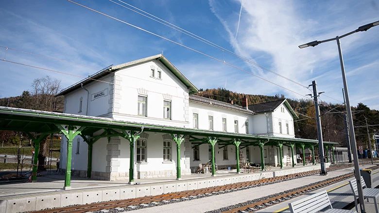 Außenansicht des Bahnhofs Mariazell mit historischem Bahnhofsgebäude, grünen Bahnsteigdächern und Gleisanlagen im Vordergrund. Der Bahnhof liegt vor einer bewaldeten Hügellandschaft unter blauem Himmel.