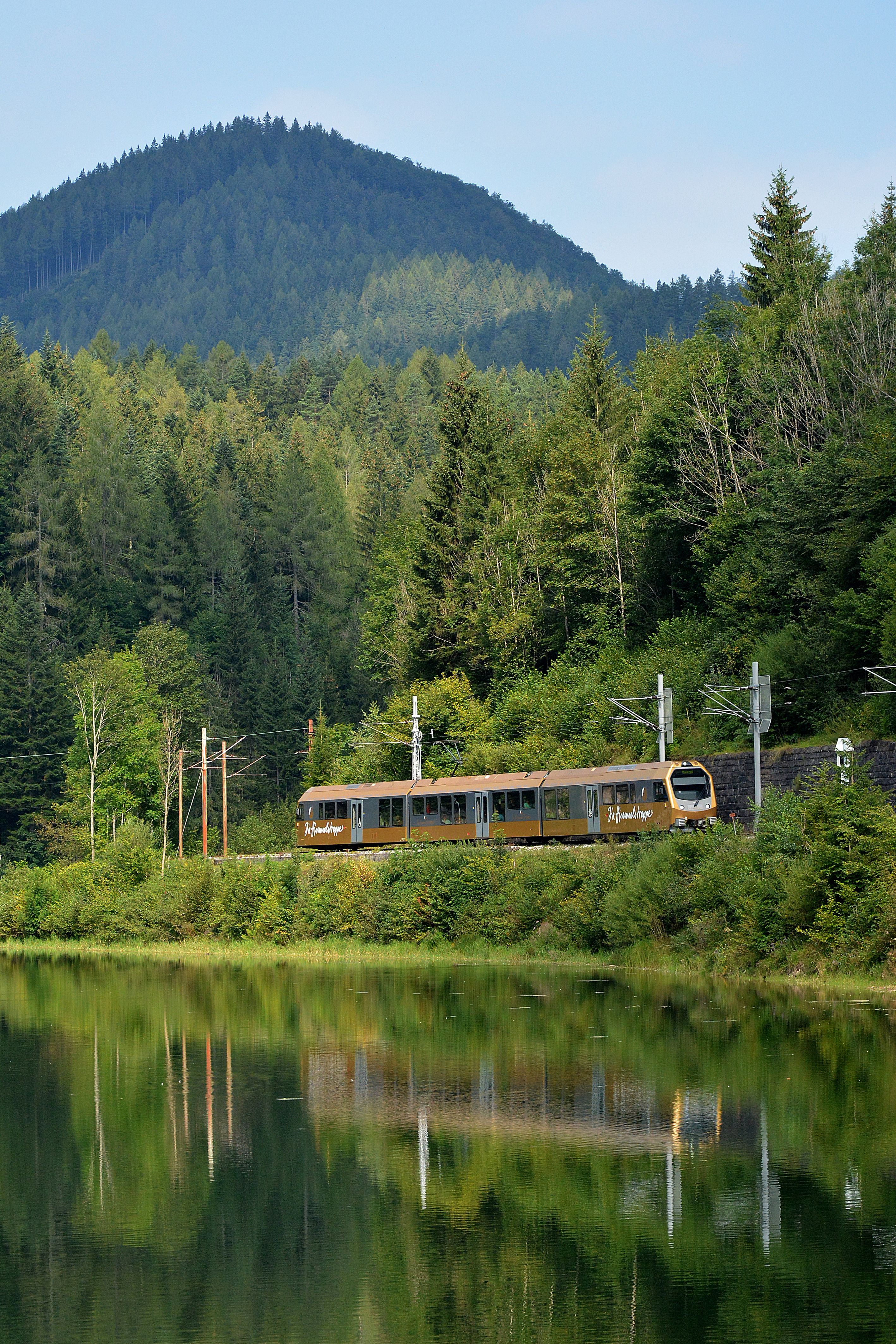 Die Himmelstreppe fährt an einem bewaldeten Hang entlang. Im Vordergrund spiegelt sich die Landschaft im Wasser, im Hintergrund ragt ein bewaldeter Berg auf.