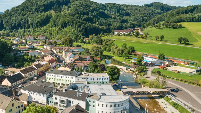 Luftaufnahme eines kleinen Dorfes mit Fluss, Brücke und umliegenden Hügeln.
