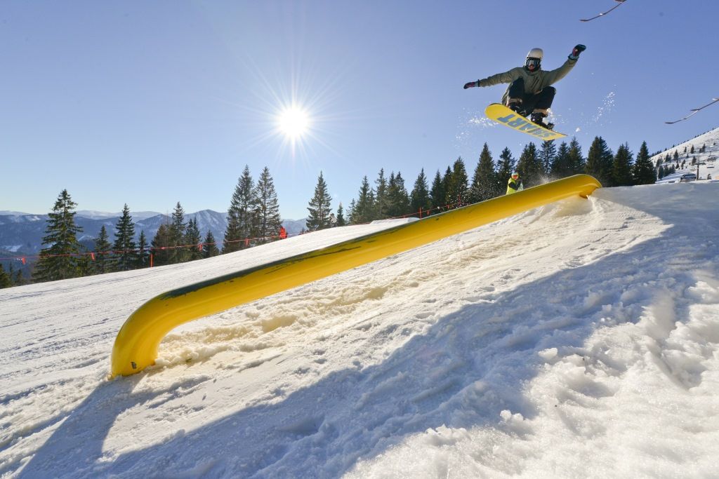 Snowboarder springt über ein gelbes Hindernis auf einer verschneiten Piste bei Sonnenschein.