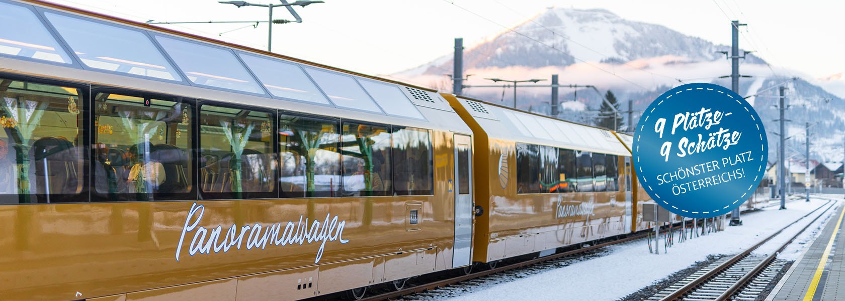 Zug mit gelb-wei&szlig;er Lackierung und der Aufschrift 'Panoramawagen' steht auf einem Bahnhofsgleis vor schneebedeckten Bergen