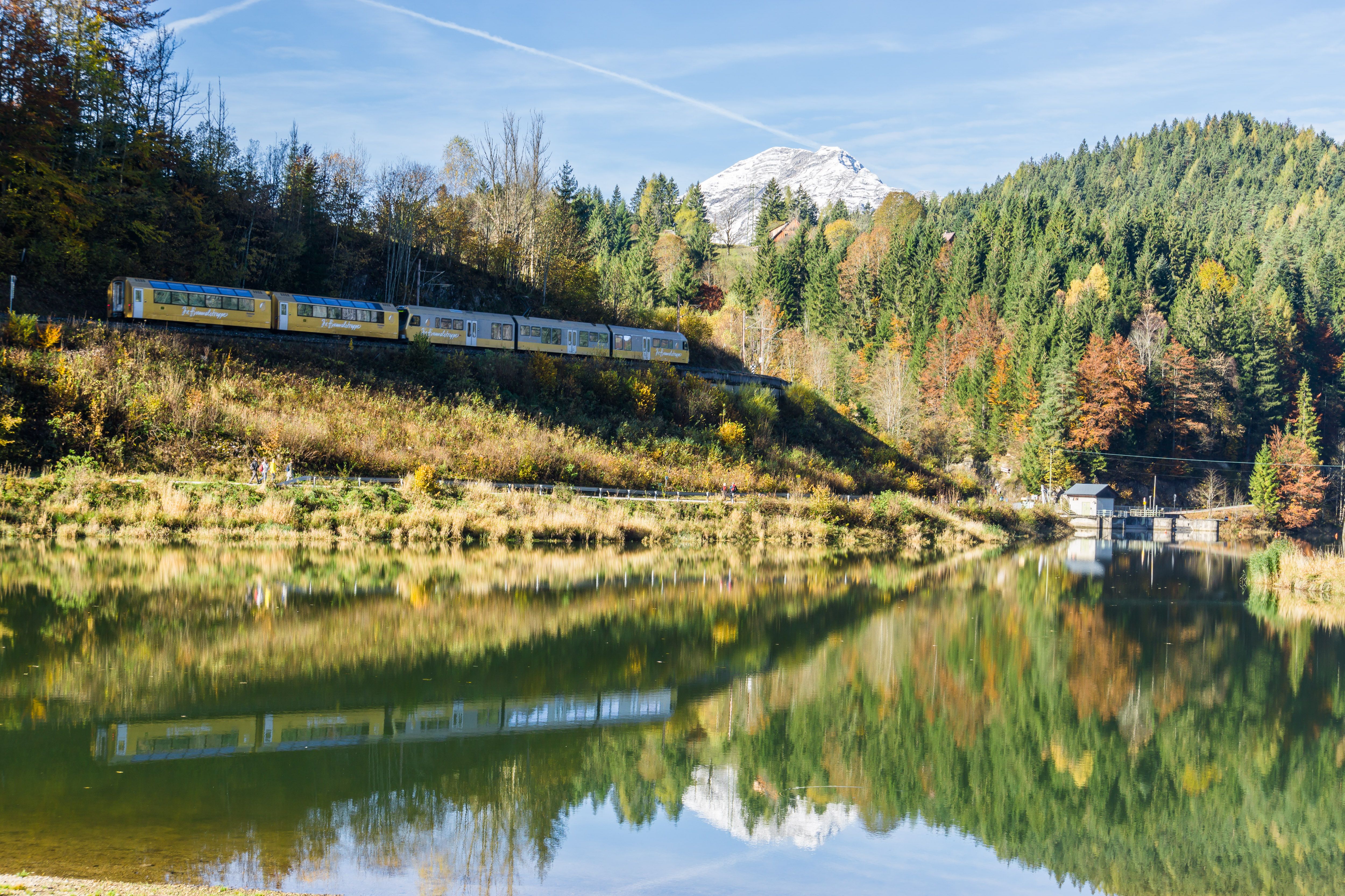 Zug fährt entlang eines bewaldeten Hangs am Ufer eines ruhigen Stausees mit Berg im Hintergrund