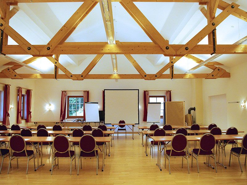 Seminarraum mit Holzbalkendecke, Tischen und Stühlen, Leinwand und Flipcharts.