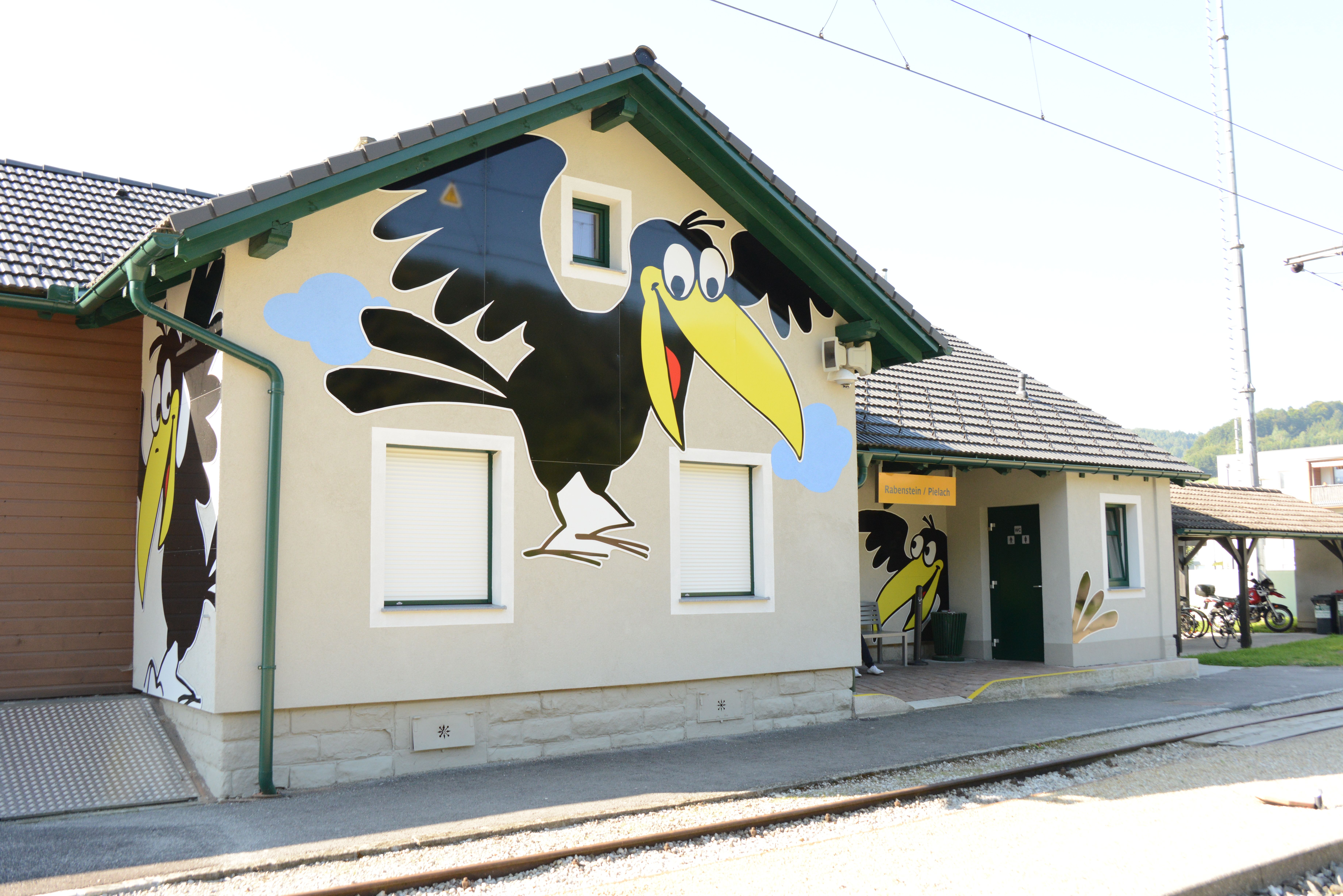 Außenansicht des Bahnhofs Rabenstein an der Pielach mit auffälliger Fassadenbemalung eines großen schwarzen Raben. Im Vordergrund verlaufen die Gleise, das Bahnhofsgebäude ist bei sonnigem Wetter zu sehen.