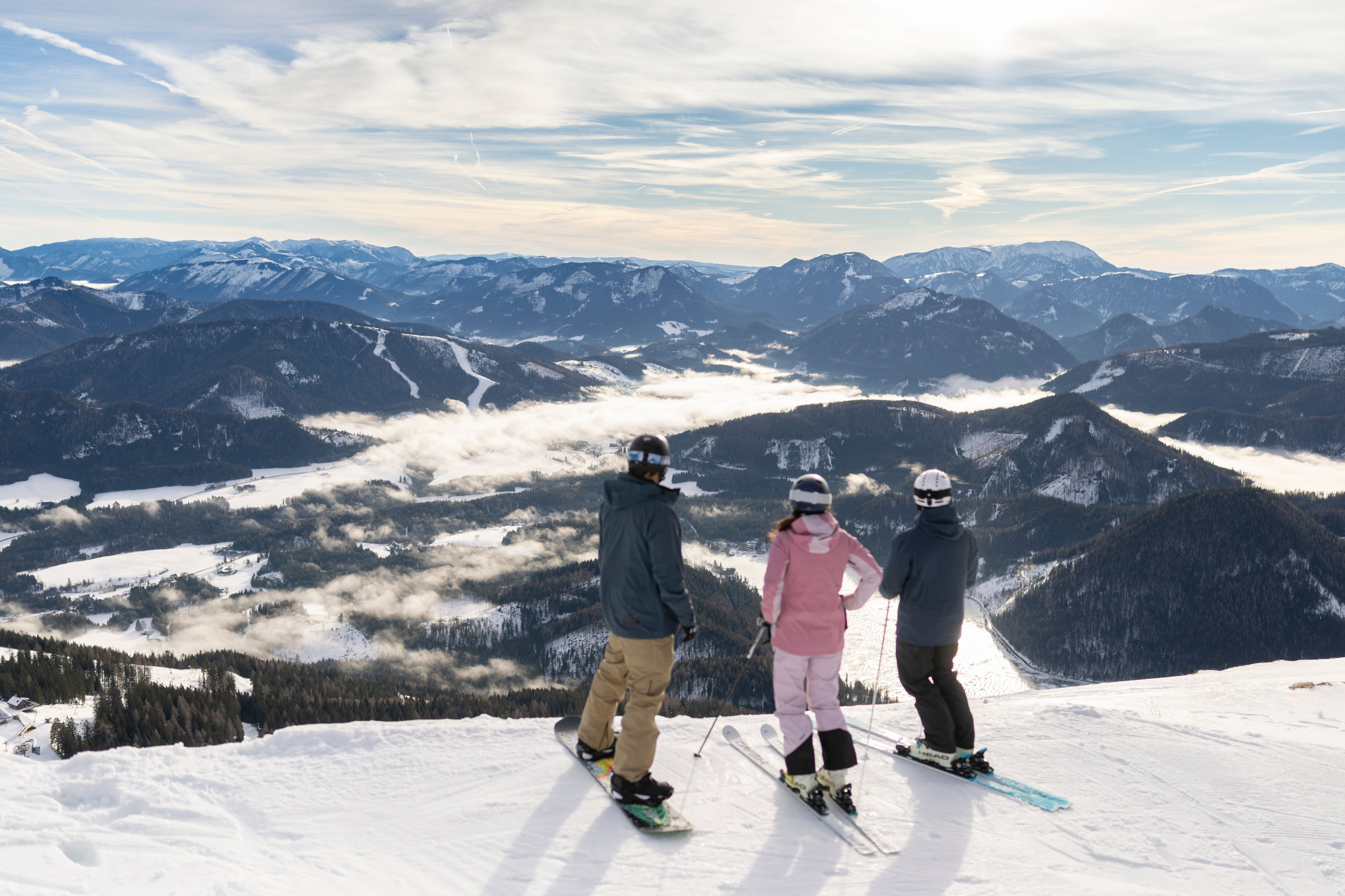 Drei Wintersportler:innen blicken von einer verschneiten Piste auf die umliegende Berglandschaft und die Nebelfelder im Tal.