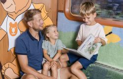 Ein Mann und zwei Kinder sitzen zusammen in einer Sitznische des Zuges. Der Junge hält ein aufgeschlagenes Märchenbuch in den Händen, während alle lächeln.