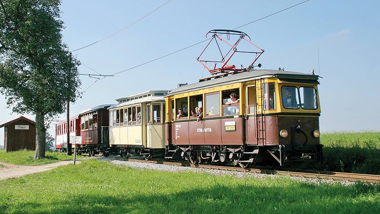 Historischer braun-gelber Triebwagen mit mehreren Waggons fährt durch eine grüne Landschaft. Fahrgäste blicken aus den offenen Fenstern des Zuges.