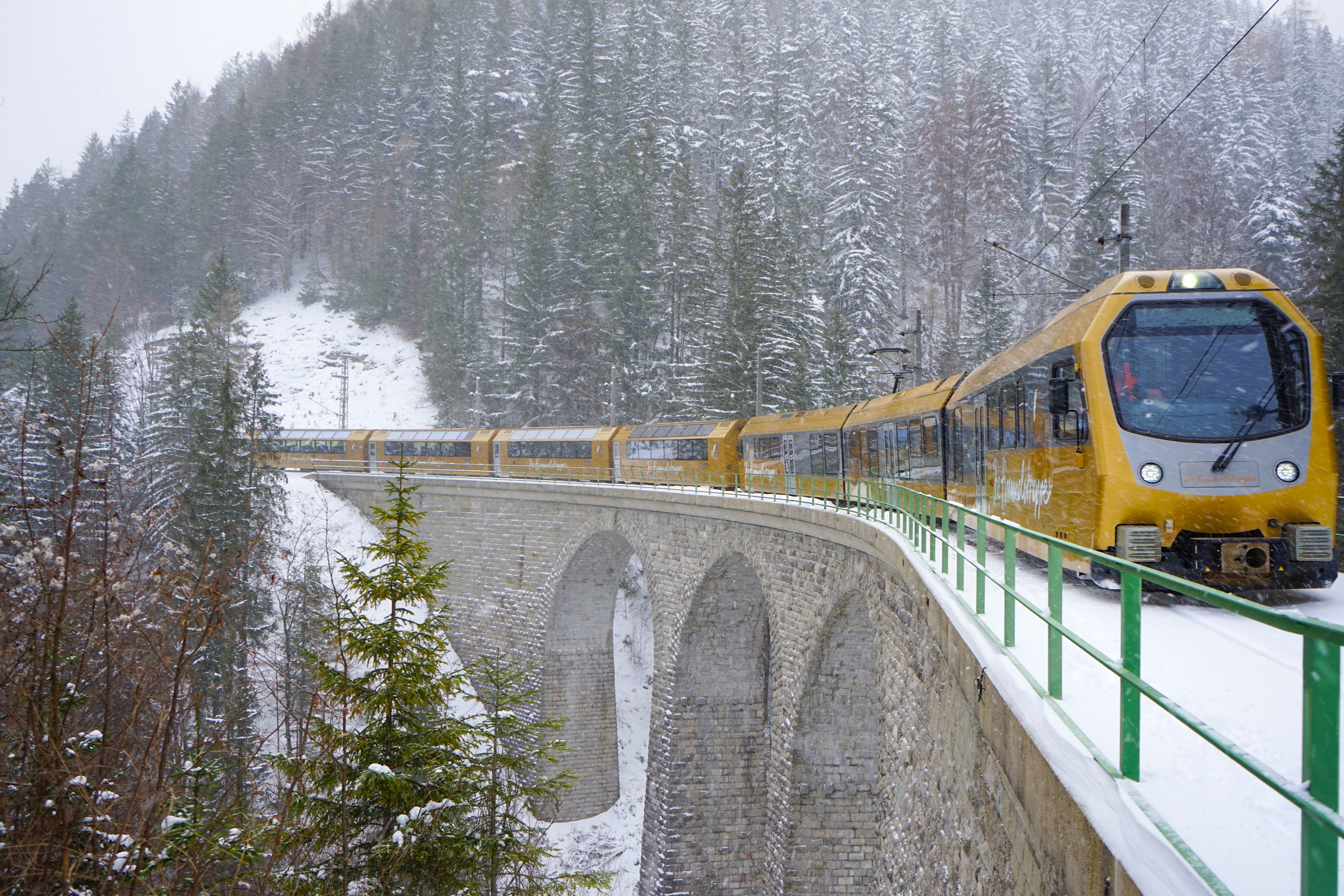 Ein gelber Panoramazug der Mariazellerbahn fährt über eine geschwungene Steinbogenbrücke durch eine verschneite Berglandschaft mit dichtem Nadelwald und schneebedeckten Hängen.