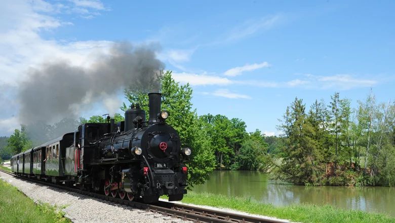 Schwarze Dampflokomotive mit grünen Waggons fährt an einem Gewässer entlang. Aus dem Schornstein steigt dichter Rauch auf; im Hintergrund stehen Bäume.