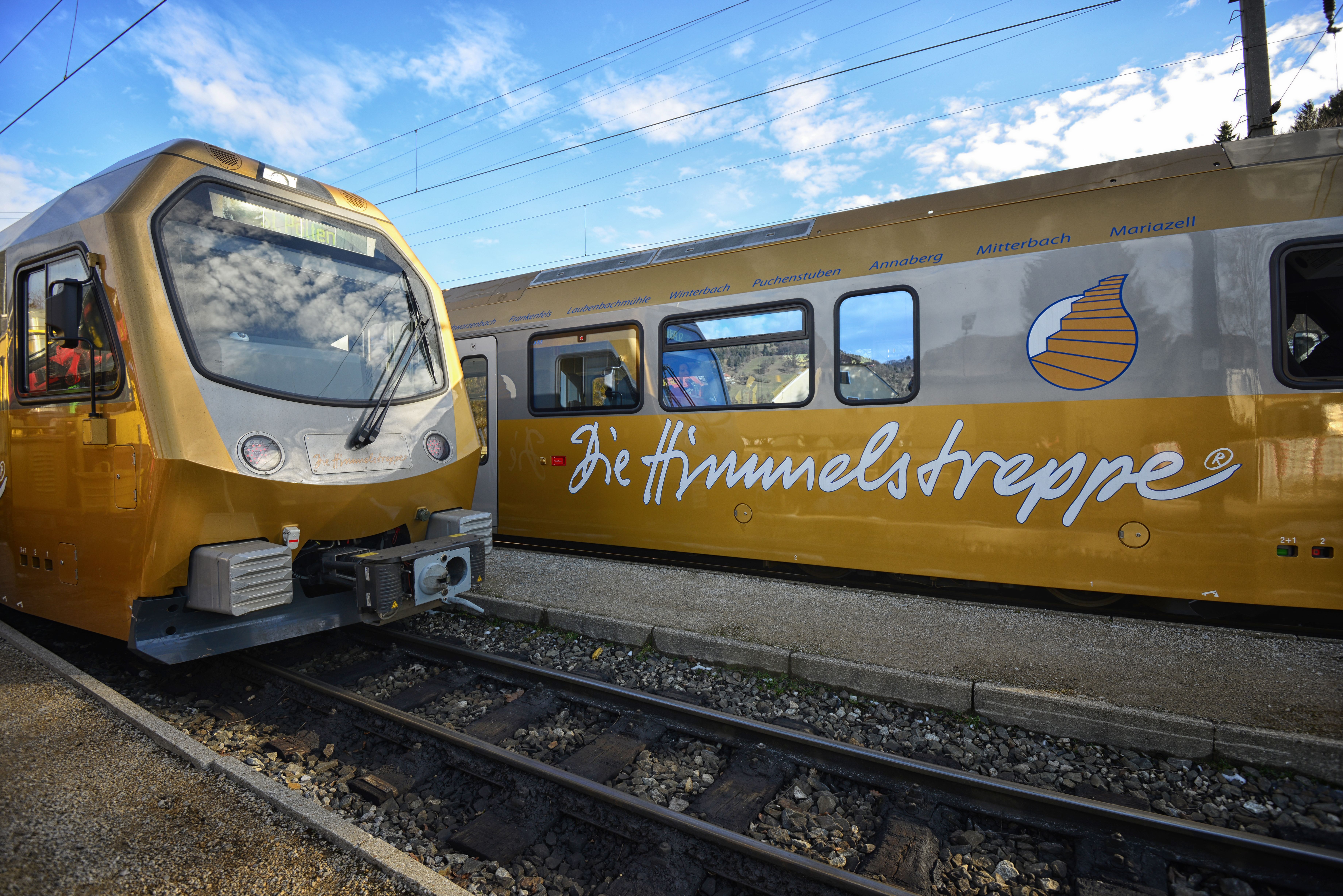 Zwei goldfarbene Züge stehen auf benachbarten Gleisen. Auf dem rechten Zug ist der Schriftzug „Die Himmelstreppe“ zu sehen, darüber sind mehrere Stationsnamen angebracht.