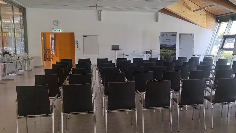 Veranstaltungsraum mit mehreren Reihen schwarzer Stühle und Präsentationstechnik.