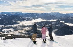 Drei Wintersportler:innen blicken von einer verschneiten Piste auf die umliegende Berglandschaft und die Nebelfelder im Tal.