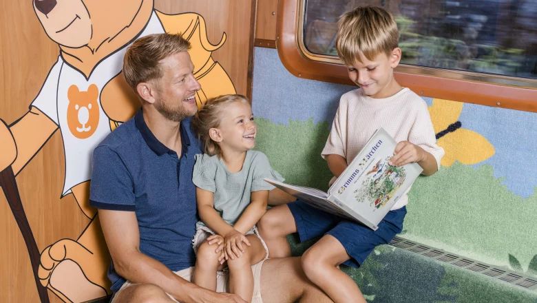 Ein Mann und zwei Kinder sitzen zusammen in einer Sitznische des Zuges. Der Junge hält ein aufgeschlagenes Märchenbuch in den Händen, während alle lächeln.