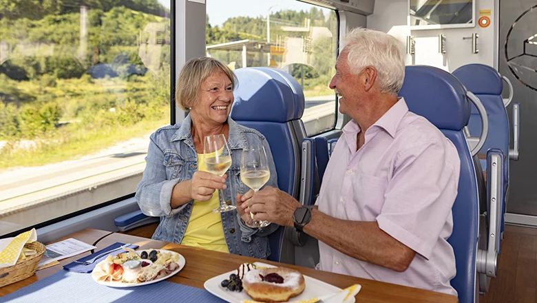Eine ältere Frau und ein älterer Mann stoßen im Zug mit Weißweingläsern über einem gedeckten Tisch an.
