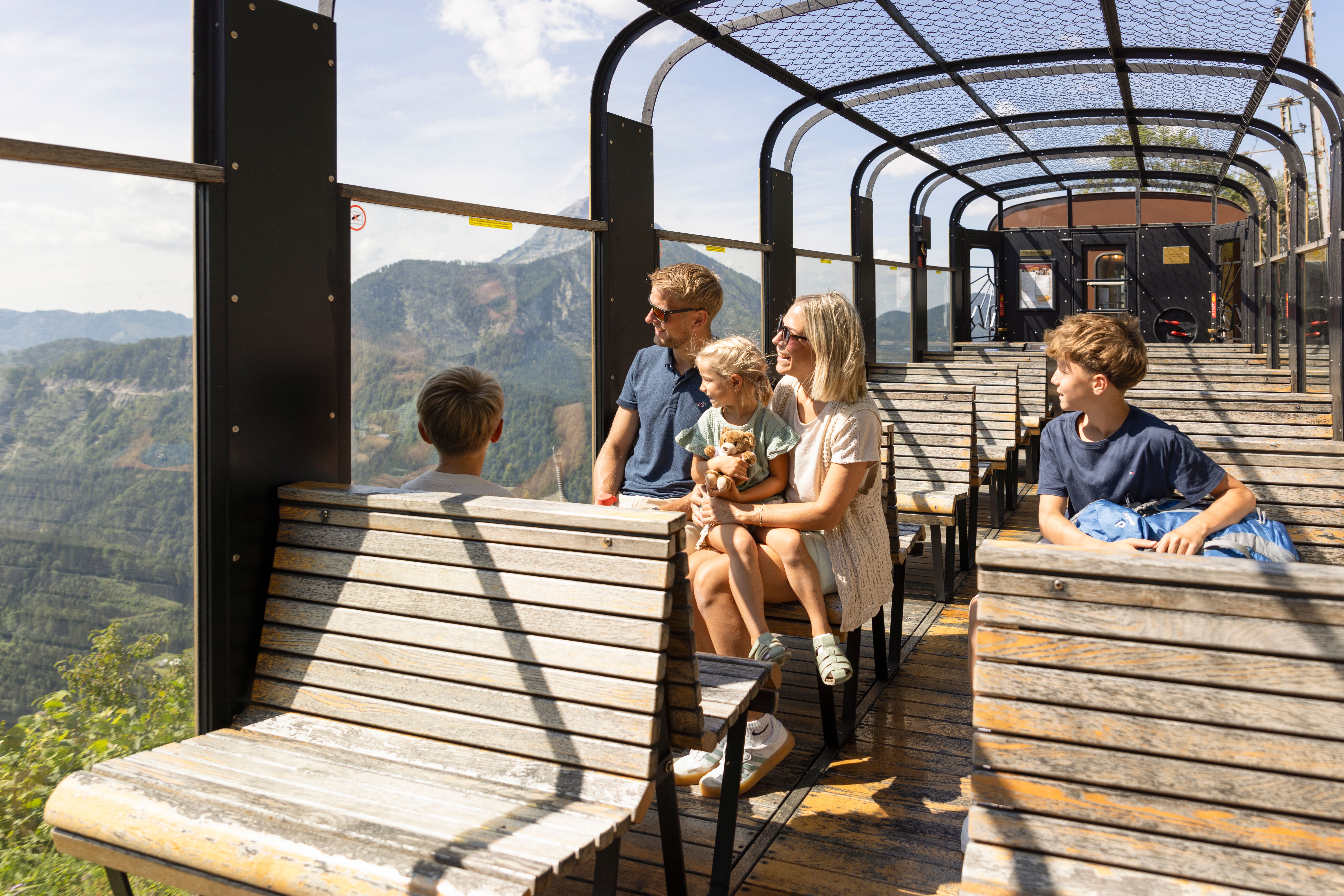 Eine Familie sitzt auf Holzbauteilen in einem offenen Aussichtswagen eines Zuges und schaut auf eine bergige Waldlandschaft unter sonnigem Himmel.