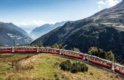 Ein rot-weißer Panoramazug fährt auf einer kurvigen Bahnstrecke durch eine eindrucksvolle alpine Berglandschaft mit weitem Blick ins Tal.
