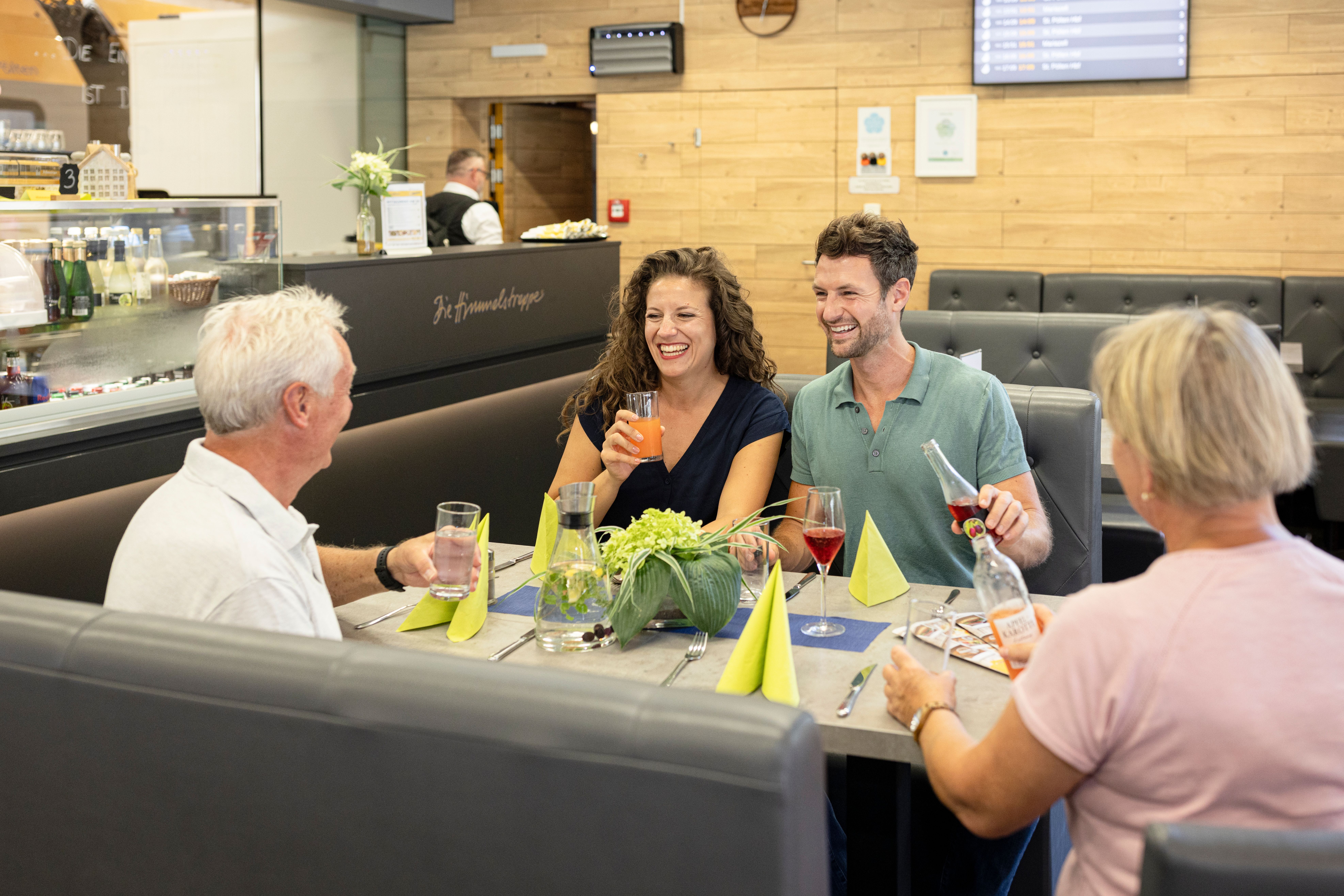 Vier Personen sitzen an einem Tisch in einem Restaurant. Zwei Personen halten Getränke in der Hand, auf dem Tisch stehen Gläser, Karaffe, Flaschen und eine Blumendekoration.