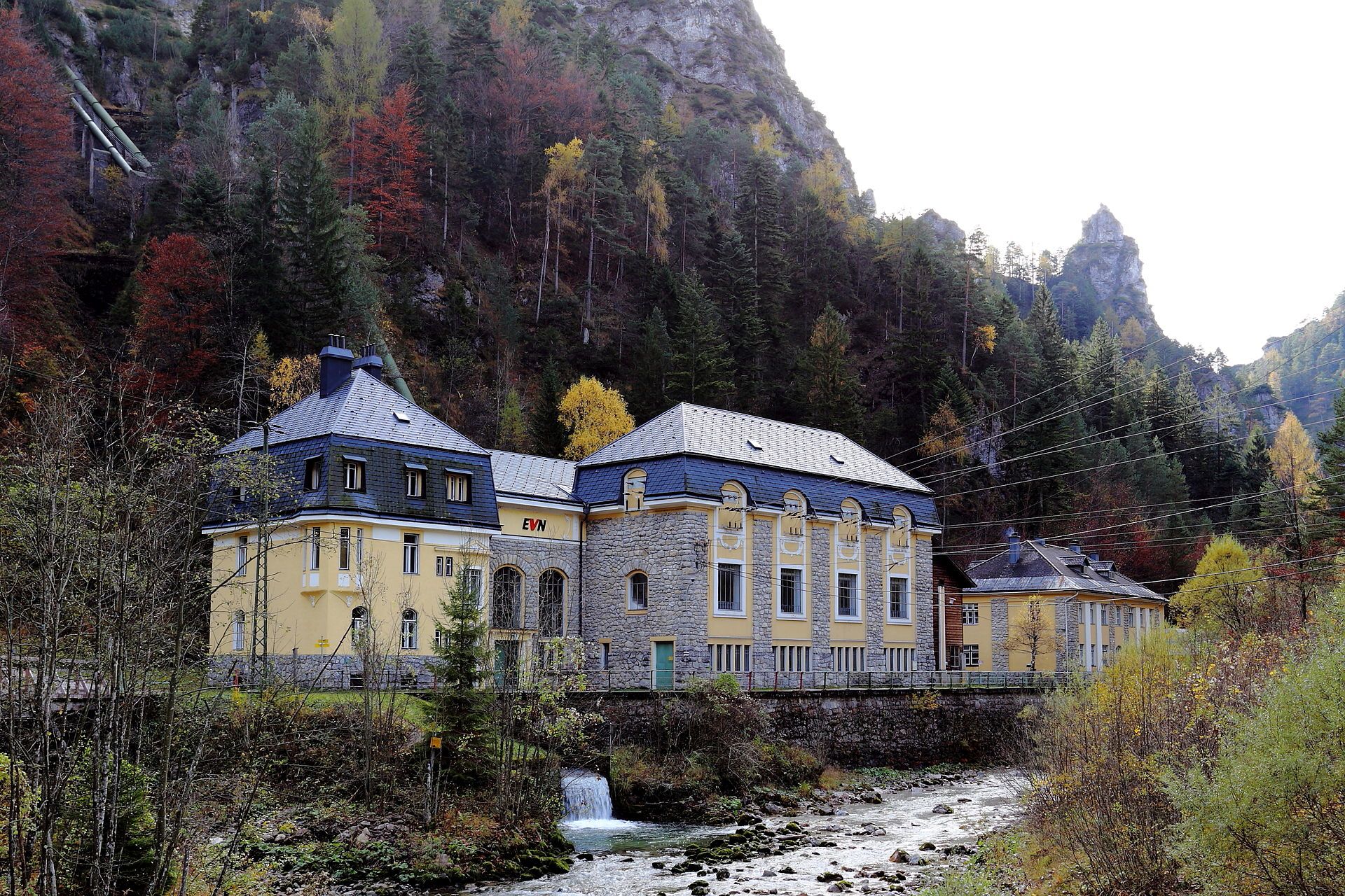 Kraftwerk Wienerbruck in bewaldeter Berglandschaft im Herbst, mit historischem Gebäude am Fluss, Stromleitungen und steilen Felsformationen im Hintergrund.