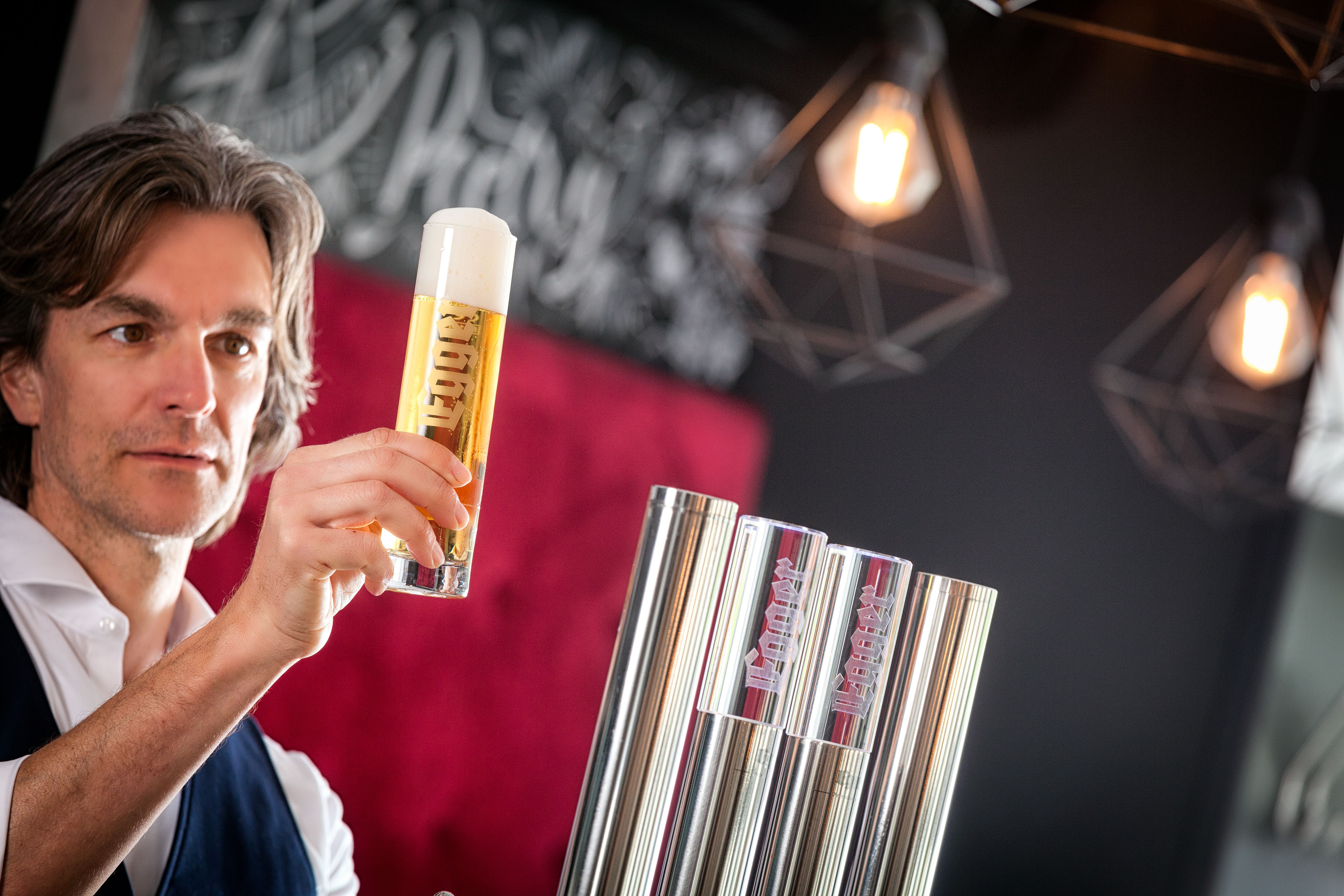 Mann hält ein Glas Bier vor einem Zapfhahn in einer Bar mit rotem Polstersofa und hängenden Lampen