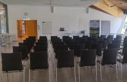 Veranstaltungsraum mit mehreren Reihen schwarzer Stühle und Präsentationstechnik.