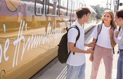 Drei junge Menschen mit Rucksäcken stehen auf einem Bahnsteig vor einem Zug mit dem Schriftzug „Die Himmelstreppe“ und unterhalten sich.