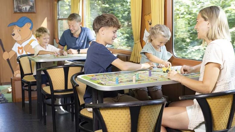 Kinder und Erwachsene sitzen im Spielewagen eines Zuges an Tischen und spielen Gesellschaftsspiele mit Spielfiguren und Würfeln. An der Wand befindet sich ein Bärenmotiv.