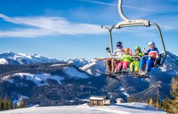 Zwei Erwachsene und zwei Kinder fahren mit Ski und Snowboards im Sessellift vor verschneiter Bergkulisse.