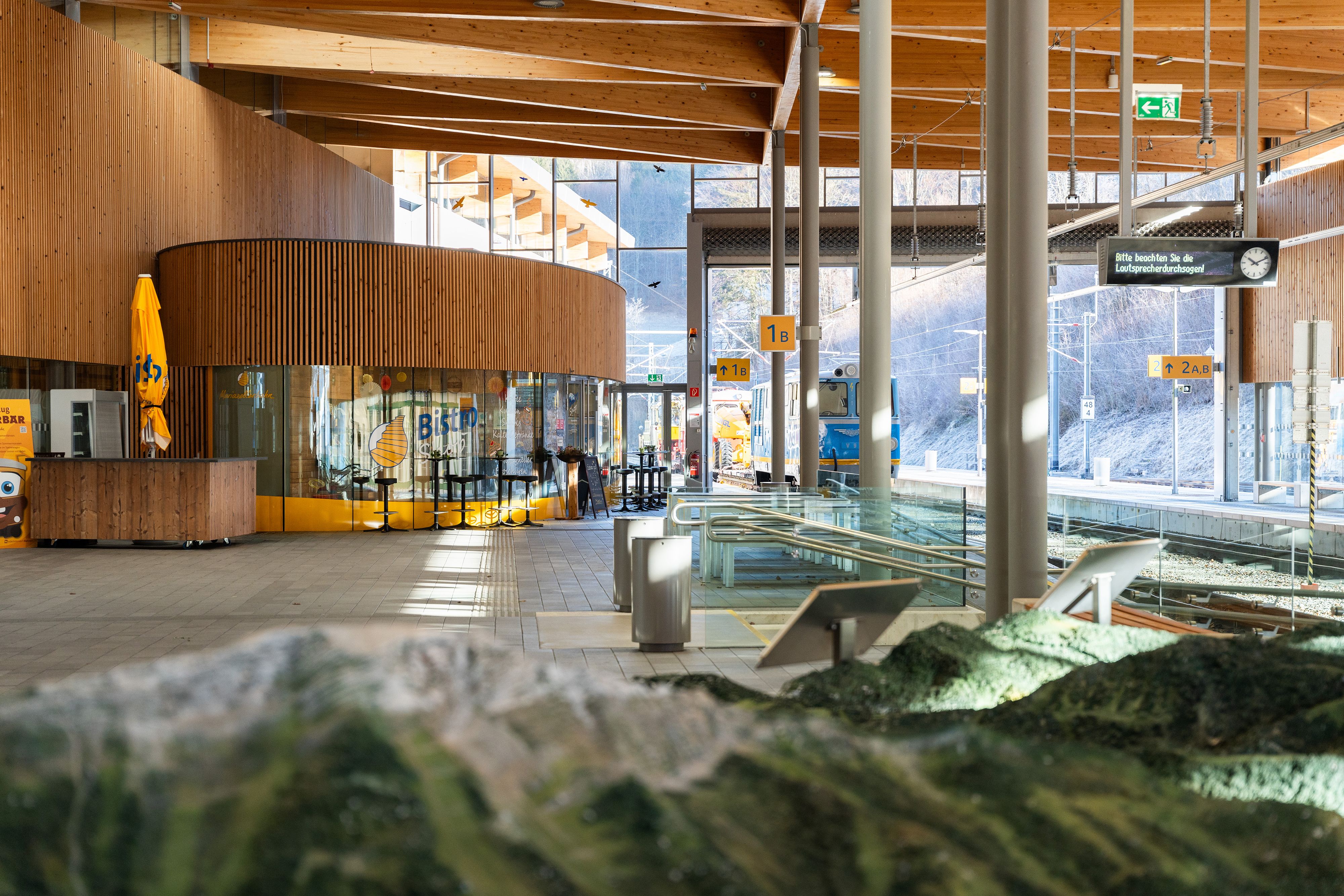 Blick in die moderne Bahnhofshalle Laubenbachmühle mit markanter Holzarchitektur, einem verglasten Bistro und einem Zug am Bahnsteig. Im Vordergrund ist ein unscharfes Landschaftsmodell zu sehen, während Tageslicht durch die großen Fenster fällt.