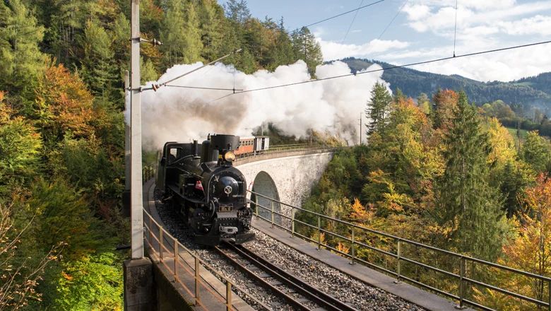 Ein Dampfzug fährt über ein steinernes Viadukt. Weiße Dampfwolken steigen in den Himmel auf, umgeben von herbstlich gefärbten Bäumen und Hügeln.