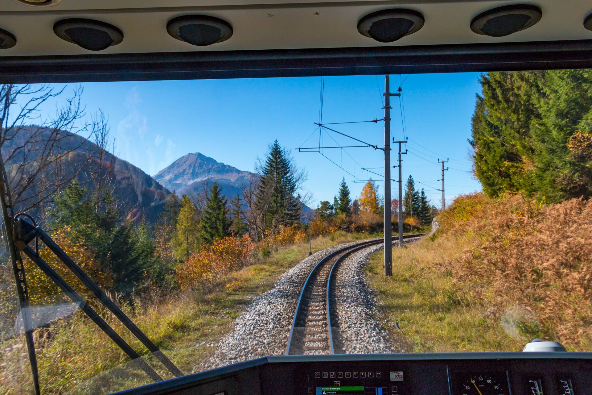 Blick aus dem Führerstand eines Zuges auf eine kurvige Bahnstrecke durch herbstliche Landschaft mit Bergen und Bäumen