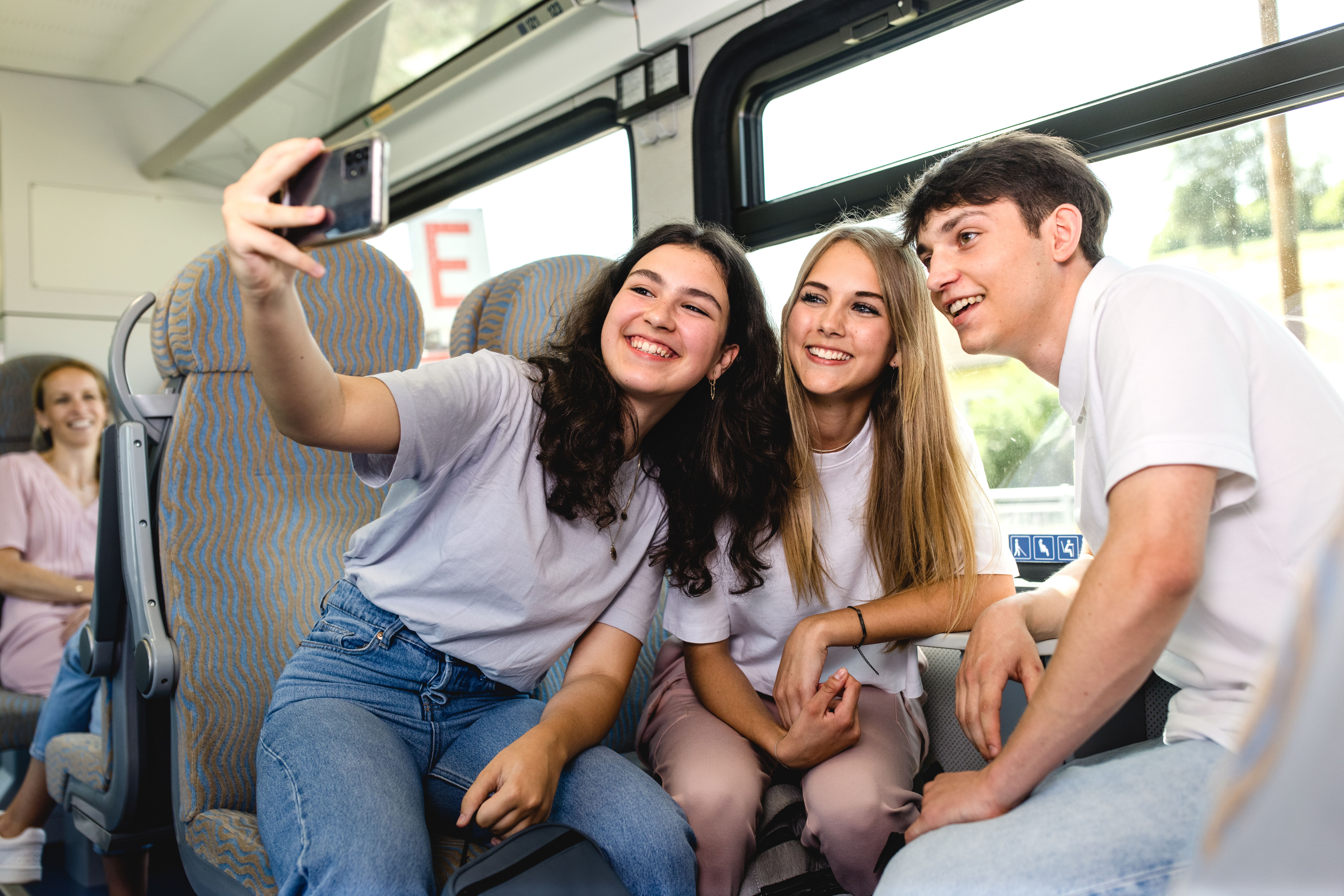 Drei junge Menschen sitzen in einem Zug, lächeln gemeinsam in die Kamera und machen ein Selfie mit dem Smartphone. Im Hintergrund sitzt eine weitere Reisende.