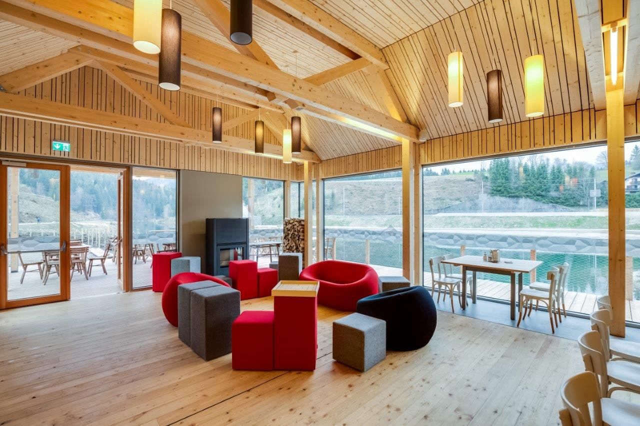 Innenansicht eines modernen Raumes mit Holzwänden, bunten Sitzmöbeln und großen Fenstern mit Blick auf eine Terrasse und Natur.