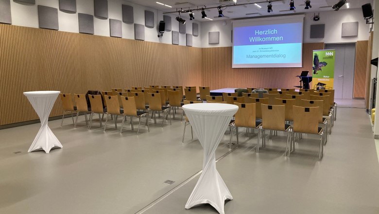 Konferenzraum mit Stuhlreihen, Stehtischen und einer Leinwand mit der Aufschrift 'Herzlich Willkommen'.