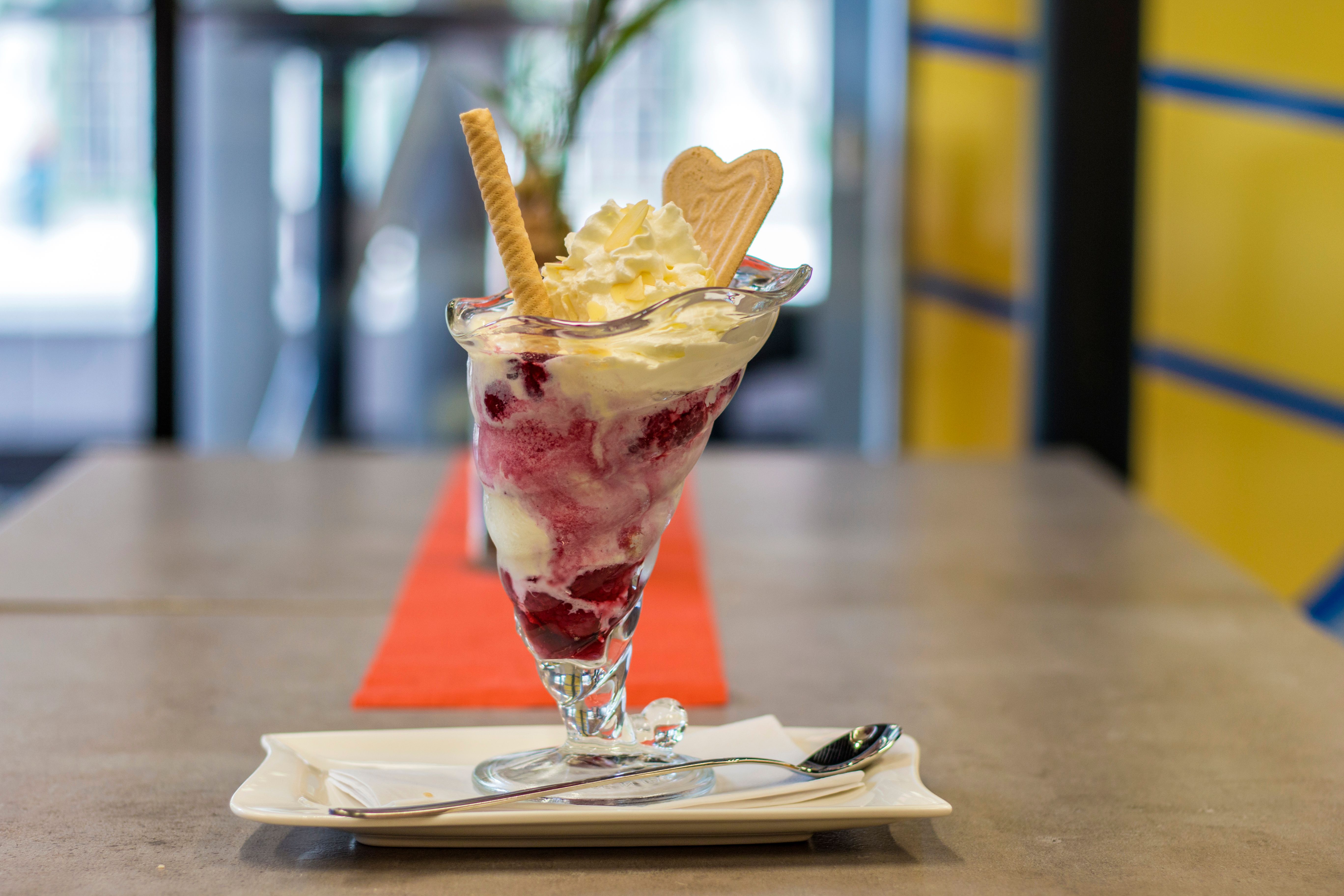 Eisbecher mit Vanilleeis, roter Fruchtsauce, Sahne, Waffelröllchen und Herzwaffel auf weißem Teller mit Löffel auf Tisch