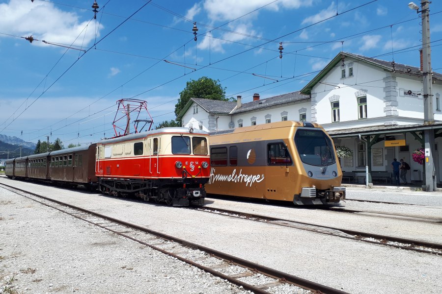 Zwei Z&uuml;ge nebeneinander an einem Bahnhof, links ein roter historischer Zug mit mehreren Waggons, rechts ein moderner goldener Zug mit der Aufschrift 'Himmelstreppe'