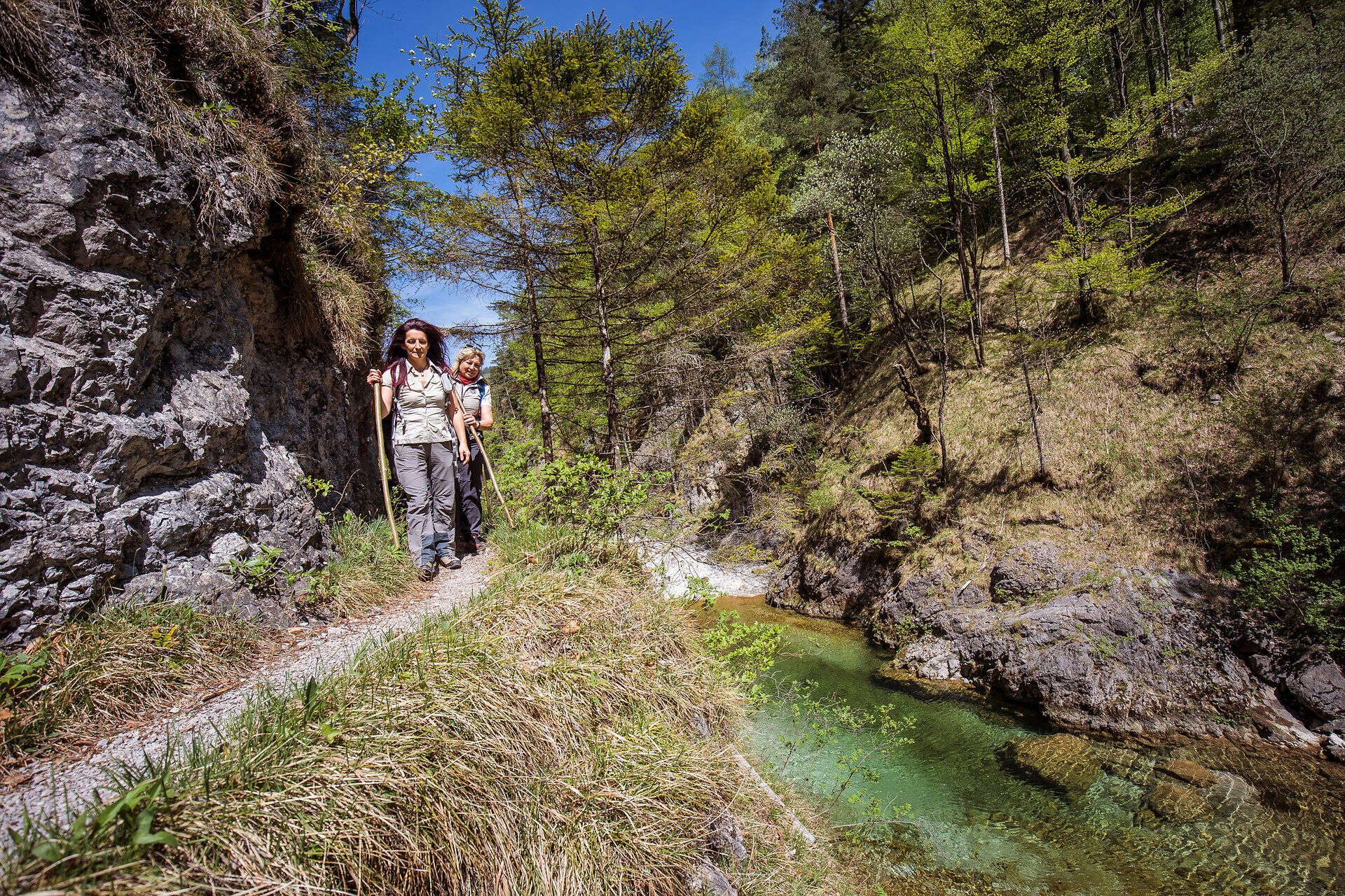 Inmitten der beeindruckenden Natur des Ötschergräben wandern zwei Abenteurer auf einem schmalen Pfad, umgeben von üppigem Grün und glitzerndem Wasser. Die sanften Hügel und die klare Luft laden dazu ein, die Schönheit der alpinen Landschaft in vollen Zügen zu genießen.