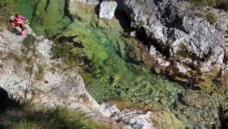 Zwei Personen sitzen auf einem Felsen neben einem klaren, gr&uuml;nen Fluss in einer Schlucht.