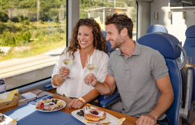Paar sitzt im Zug und st&ouml;&szlig;t mit Wei&szlig;wein an, Jause steht auf dem Tisch.