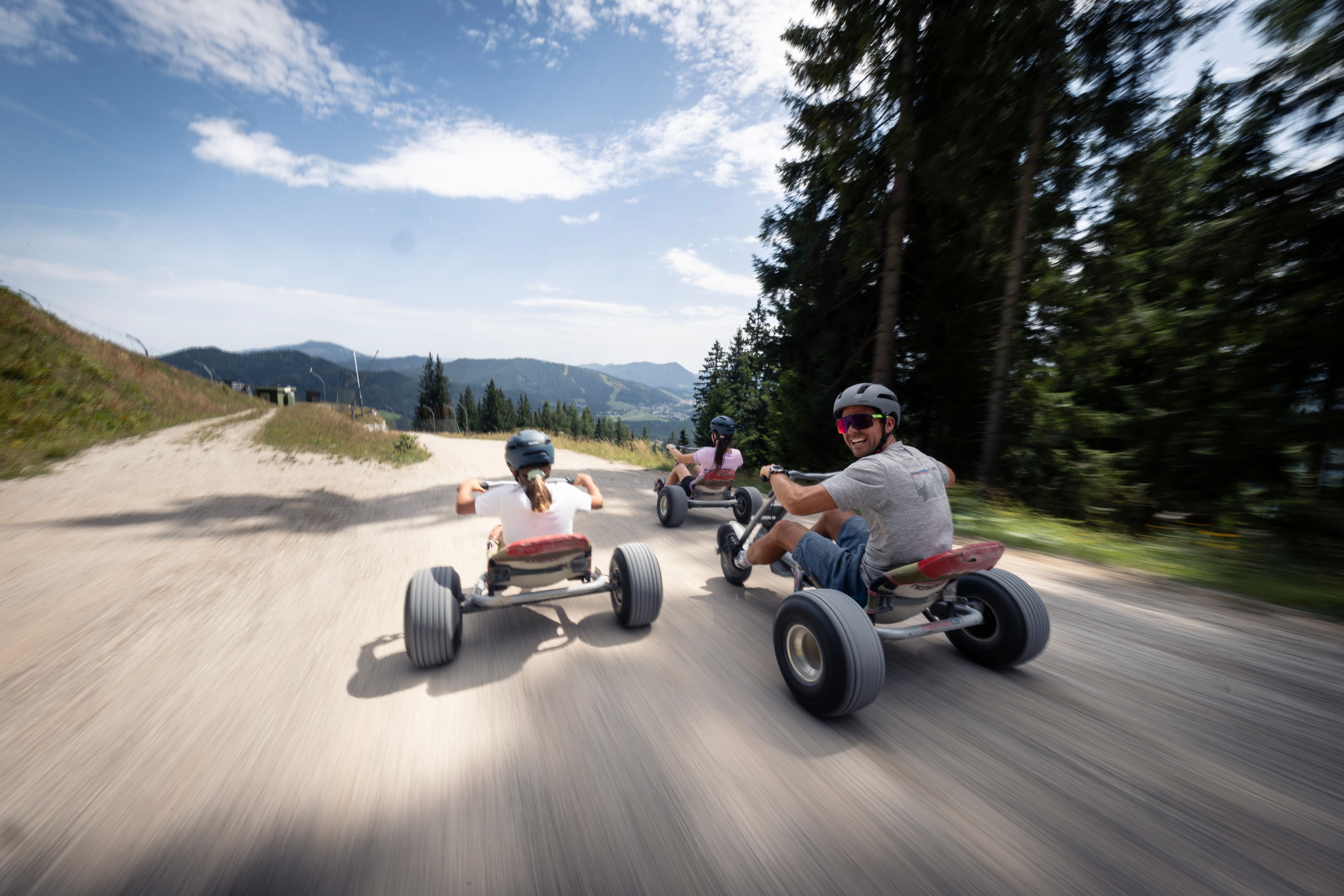 Familie fährt mit Mountaincarts rasant talwärts; der Vater blickt lachend zurück.