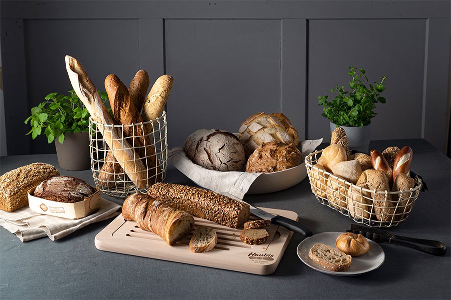 Verschiedene Brotsorten wie Baguette, Körnerbrot und Brötchen in Drahtkörben und auf Holzbrett mit Kräutertöpfen im Hintergrund