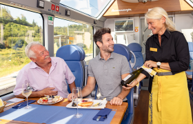 Schaffnerin serviert Wein im Panoramawagen, Fahrg&auml;ste genie&szlig;en Essen und Aussicht.