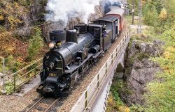 Die schwarze Dampflokomotive Mh.6 fährt mit ihren Waggons über eine Brücke. Dahinter führt ein Gleis aus einem Tunnel in einer felsigen Waldlandschaft.